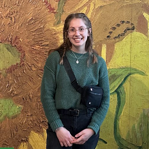 girl with brown hair in braids, round glasses, green sweater, in front of van gogh's sunflowers painting